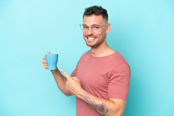 Jeune homme caucasien tenant une tasse de café isolé sur fond bleu et le pointant - Photo, image