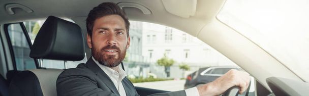 Handsome businessman in grey suit is riding behind steering wheel of car. Blurred background - 写真・画像