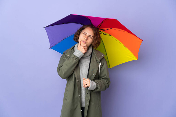 English woman holding an umbrella isolated on purple background and looking up - Photo, Image