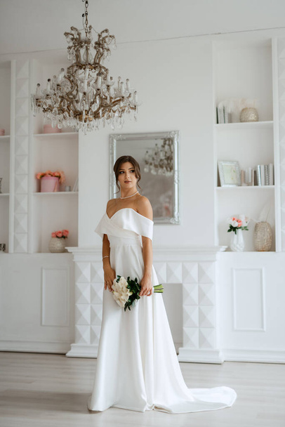 brunette bruid in een strakke bruiloft jurk in een heldere studio met een boeket - Foto, afbeelding