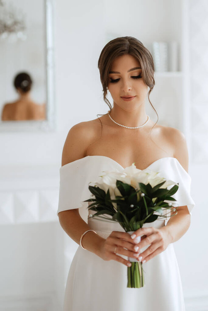 brunette bride in a tight wedding dress in a bright studio with a bouquet - Фото, изображение