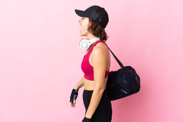 Молодая спортсменка со спортивной сумкой на изолированном фоне смеется в боковом положении - Фото, изображение