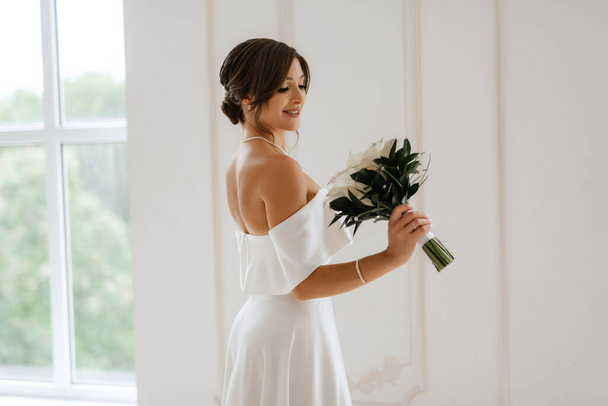 brunette bride in a tight wedding dress in a bright studio with a bouquet - Fotó, kép