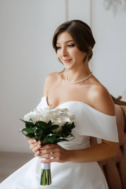 brunette bride in a tight wedding dress in a bright studio with a bouquet on an armchair - Photo, Image