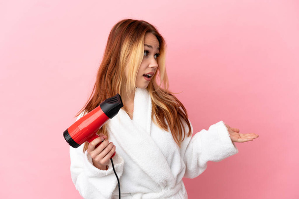 Teenager blonde girl holding a hairdryer over isolated pink background with surprise expression while looking side - Φωτογραφία, εικόνα