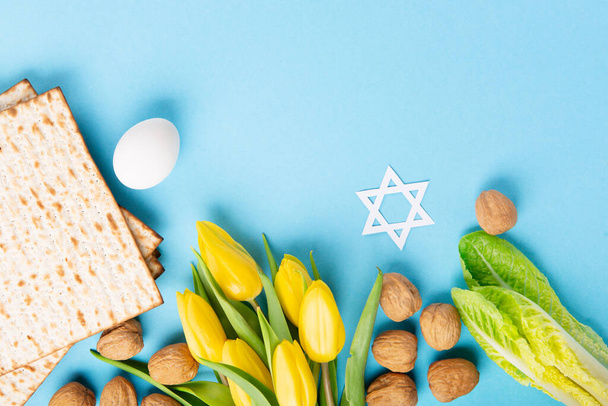 Jewish holiday Passover greeting card concept with matzah matzoh (jewish holiday bread), walnuts, yellow tulip flowers on blue table. Seder Pesach spring holiday background, copy space. - Фото, изображение