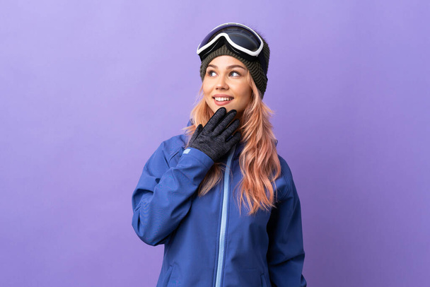 Skier teenager girl with snowboarding glasses over isolated purple background looking up while smiling - Photo, Image