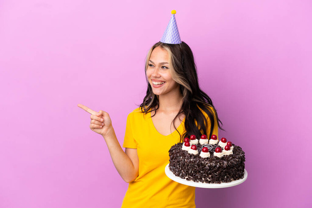 Adolescente russe fille tenant gâteau d'anniversaire isolé sur fond violet pointant du doigt sur le côté et présentant un produit - Photo, image