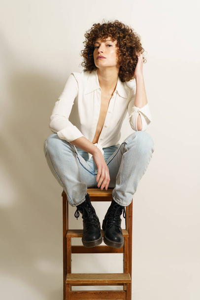 Full length of flirty woman model with curly hair, in stylish wear resting on wooden stool playing with curl and looking at camera against white background - Foto, afbeelding