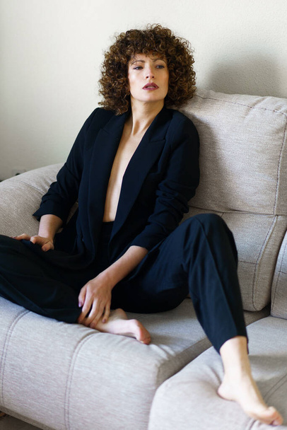 Cuerpo completo de alto ángulo de la mujer adulta con el pelo rizado oscuro sentado en el sofá con chaqueta negra y pantalones mirando a la cámara - Foto, Imagen