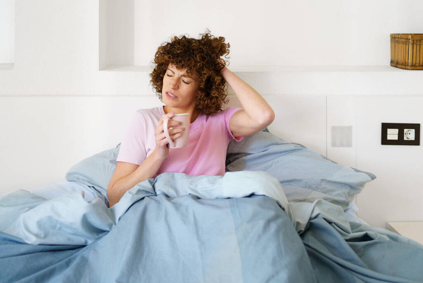 Lady touching head with closed eyes while sitting on bed and drinking hot beverage in morning - Photo, Image