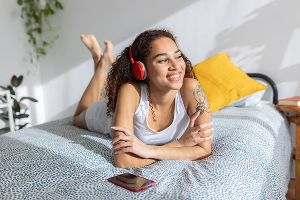 Donna latinoamericana godendo di musica online al telefono. Podcast d'ascolto femminile millenario sul letto. Intrattenimento e concetto di persone - Foto, immagini