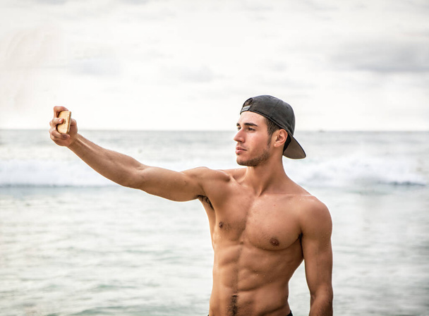 Metade do corpo de um jovem bonito usando o celular para tirar foto, de pé em uma praia, sem camisa vestindo shorts boxeador e chapéu de beisebol, mostrando corpo musculoso - Foto, Imagem