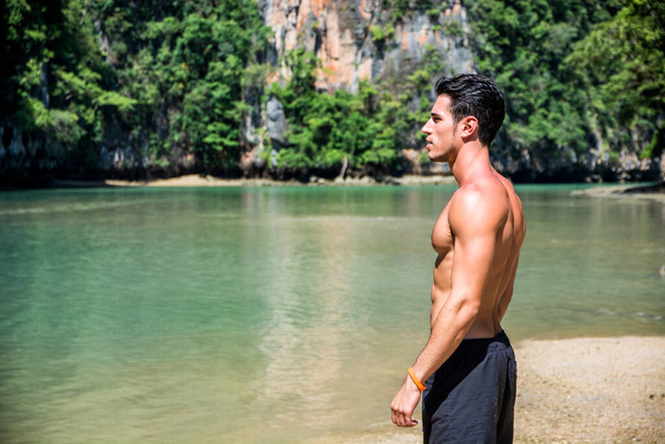 Metade do corpo de um jovem bonito em pé em uma praia em Phuket Island, Tailândia, sem camisa vestindo shorts boxer, mostrando corpo musculoso em forma - Foto, Imagem