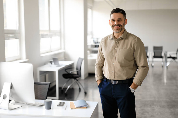 Successful person. Portrait of happy middle aged businessman in shirt posing with hands in pockets and smiling, office interior, copy space, empty mockup place for advertisement - Foto, Imagen