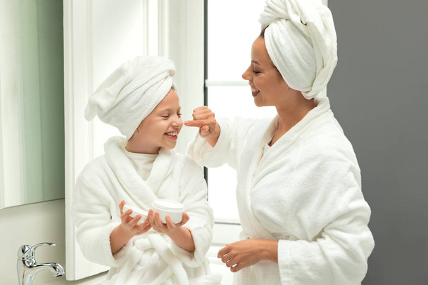 Femme millénaire européenne souriante et petite fille en peignoirs et serviettes s'amusent, prennent soin de la beauté et de la peau, appliquent de la crème sur le visage dans l'intérieur de la salle de bain blanche. Bonjour et relation - Photo, image
