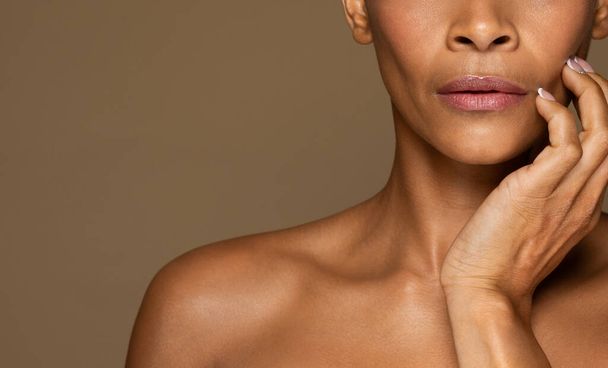 頬に触れる黒人の中年女性のクロップ,完璧な肌を示す,茶色のスタジオの背景に孤立ポーズ,自由空間.スキンケア、化粧品手順の概念 - 写真・画像