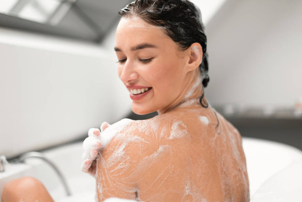 Pretty Woman mytí těla s pěnou nanášení sprchový gel na rameno sedí zpět na kameru ve vaně v moderní koupelně Indoors. Bodycare Beauty Rutinní koncepce. Selektivní zaměření - Fotografie, Obrázek