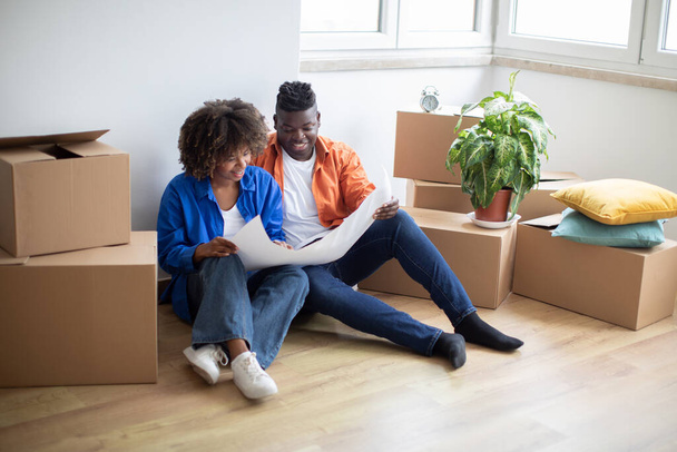 Boldog fekete házastársak, akik az új otthonba költözés után tervrajzokat nézegetnek, fiatal afro-amerikai párok, akik együtt tervezik a lakásukat, a padlón ülnek kartondobozok között az áthelyezés után. - Fotó, kép