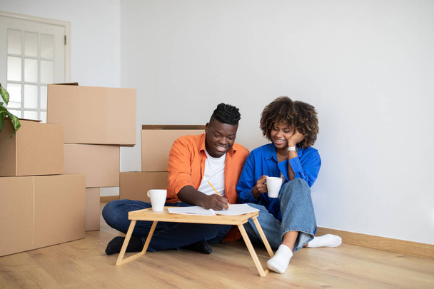 Happy Black Couple Making List Of Necessities And Drinking Coffee After Moving Home, Young African American Spouses Planning Shopping Or Calculating Budget For House Renovation, Sitting Among Boxes - Zdjęcie, obraz