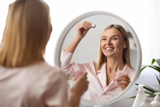 Концепция Skincare. Красивая женщина средних лет, применяя сыворотку для лица с каплей, стоя рядом с зеркалом дома, счастливая зрелая женщина увлажняющая кожу и улыбающаяся своему отражению, избирательный фокус - Фото, изображение
