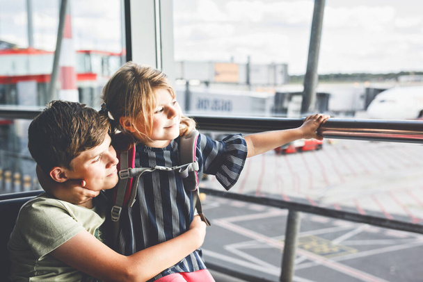 Κοριτσάκι και σχολιαρόπαιδο στο αεροδρόμιο περιμένουν επιβίβαση στο μεγάλο παράθυρο. Δύο παιδιά στέκονται στο παράθυρο με φόντο τα αεροπλάνα. Ευτυχισμένα παιδιά, αδέλφια φεύγουν για οικογενειακές καλοκαιρινές διακοπές. - Φωτογραφία, εικόνα