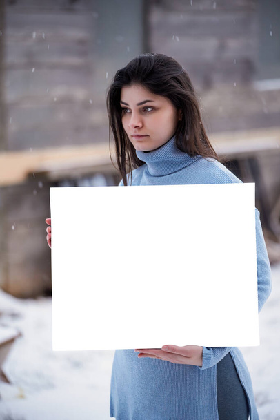 Hermosa mujer tiene una tarjeta blanca en blanco. Chica morena mientras nieve al aire libre. maqueta de arte - Foto, imagen