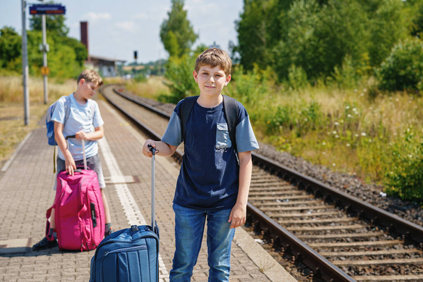 Two children, school boys with suitcases before leaving for summer vacation camp. Happy kids, siblings, twins brothers going on journey, family road trip waiting for train - Photo, image