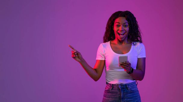 Świetna oferta. Podekscytowany młody czarny kobieta gospodarstwa smartfon i wskazując obok na przestrzeń kopiowania stojąc w Neon Light nad fioletowym tle Studio, Afroamerykanka Showign wolne miejsce na reklamę - Zdjęcie, obraz