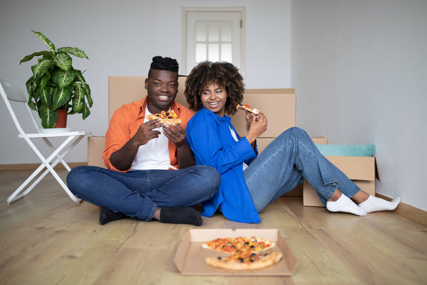 Heureux couple noir manger de la pizza tout en ayant pause le jour du déménagement, joyeux conjoints afro-américains assis sur le sol parmi les boîtes en carton, célébrer la réinstallation dans un nouvel appartement - Photo, image