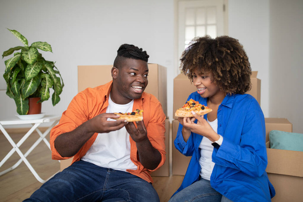 Χαρούμενο μαύρο ζευγάρι που κάνει διάλειμμα για πίτσα μετά τη μετακόμιση σε νέο σπίτι, ευτυχισμένοι νεαροί αφρικανοί σύζυγοι που κάθονται στο πάτωμα ανάμεσα σε χαρτόκουτα, γιορτάζοντας τη μετεγκατάσταση στο δικό τους σπίτι - Φωτογραφία, εικόνα