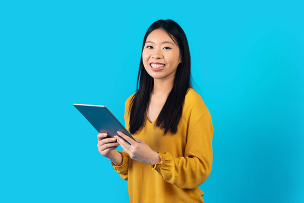 Technologies innovantes. Jeune jolie femme chinoise lisant blog ou ebook en ligne, en utilisant un pad numérique PC moderne et souriant à la caméra, isolé sur fond bleu studio, espace de copie - Photo, image