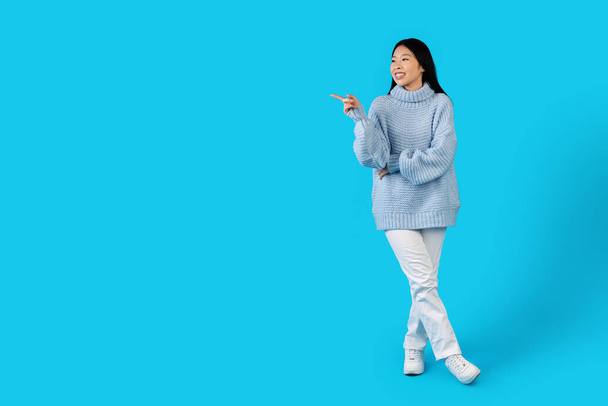 Симпатичная привлекательная длинноволосая молодая азиатка в тёплом свитере и брюках, указывающая на копировальное место для рекламы и улыбки, изолированная на синем фоне студии, полная длина - Фото, изображение