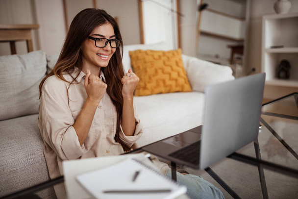 Glad smiling millennial mixed race woman in glasses has video call on laptop, making victory and success gesture in room interior. Win, great work, business remotely and study at home - Fotó, kép
