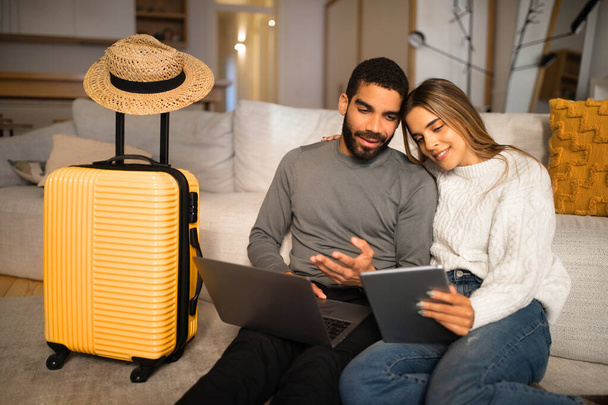 Smiling multiracial young couple sit on floor with gadgets, suitcase and hat, choose country for vacation in living room interior. Travel planning, app for prip, pay for tickets - Foto, imagen
