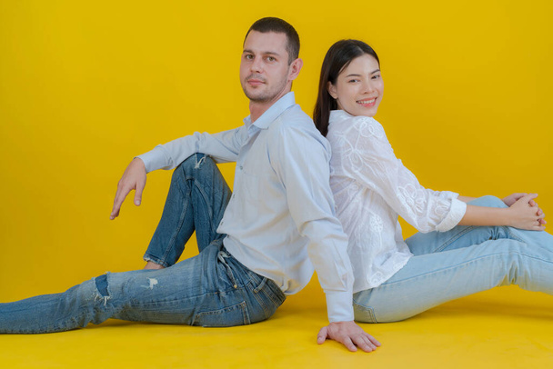Ζευγάρι χαμογελά και κάθεται μαζί στο πάτωμα και κοιτάζοντας κάμερα σε κίτρινο φόντο. - Φωτογραφία, εικόνα
