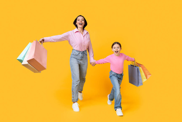 Family Shopping. Happy Mother And Little Daughter Walking With Bright Shopper Bags Over Yellow Studio Background, Cheerful Mom And Female Child Enjoying Seasonal Sales And Discounts, Copy Space - Photo, Image