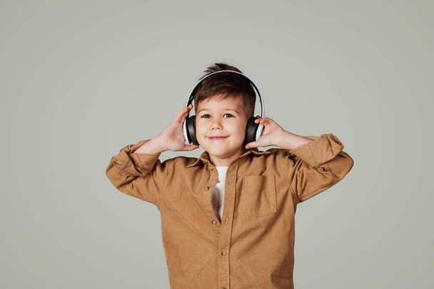 Smiling little european boy in wireless headphones listening music, enjoy free time isolated on gray studio background. Schoolchild have fun with audio app, study and education - Valokuva, kuva