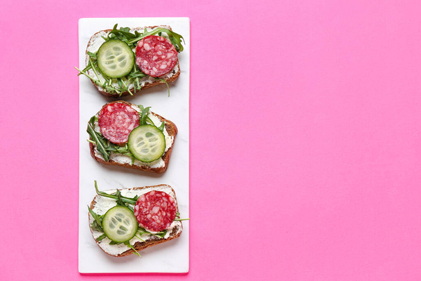 Placa de sabrosos sándwiches con queso crema, salami, pepino y rúcula sobre fondo de color - Foto, imagen