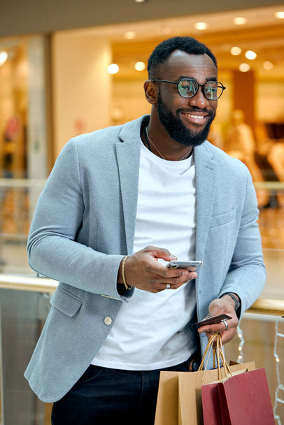 Happy smiling handsome young bearded man holding smartphone, card and colorful shopping bags.happiness, man buying goods online, close up side view photo.shopping addiction. - Fotografie, Obrázek