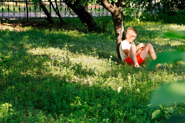Onnellinen lapsi teini poika istuu vihreällä ruoholla ulkona kevätpuistossa tai puutarhassa. Onnellinen lapsi poika niityllä kesällä luonnossa. Unelmaidea. Kopioi tila. - Valokuva, kuva