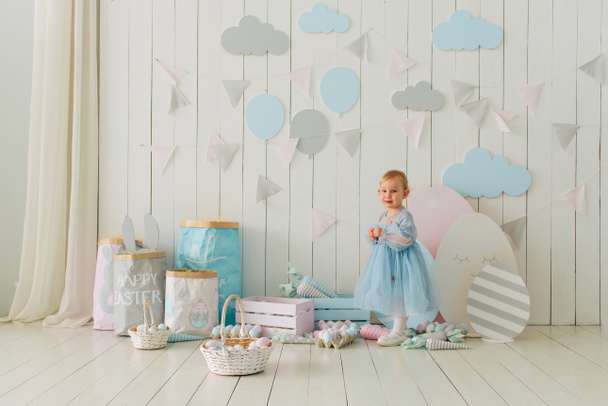 cute little girl in a smart dress is holding a basket with a decorated easter eggs. Easter holidays in decorated living room with lots of Easter eggs and decorative carrots, selective focus - Foto, Imagen