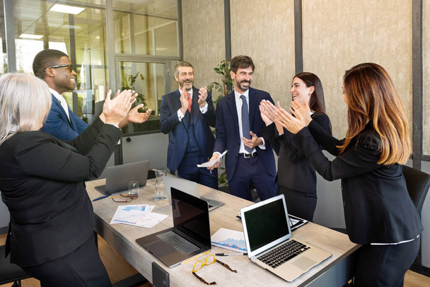 Команда мультирасовых бизнесменов в шикарных костюмах собирается за столом с ноутбуками в конференц-зале и аплодирует после успешной встречи - Фото, изображение