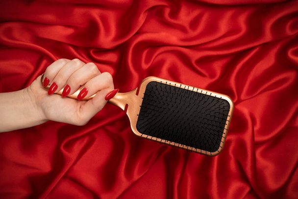 A red hairbrush in woman hand on a red silk background. Beauty tools. Hair tools.  - Foto, Imagem