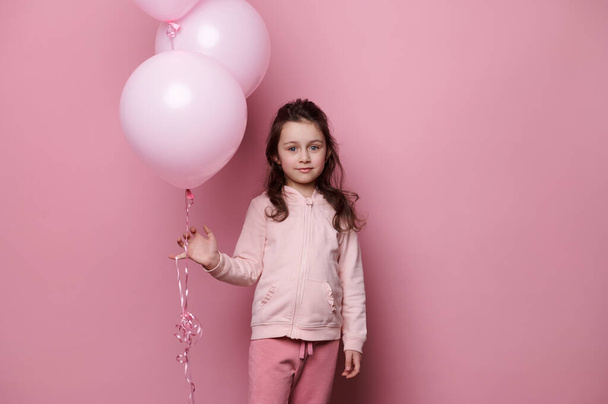 Adorable niño caucásico de 5-6 años, encantadora niña con ropa rosa, sosteniendo un montón de globos de helio pastel rosa, sonrisas, mirando a la cámara, aislado en el fondo de color rosa - Foto, Imagen