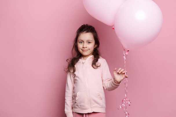 Retrato sobre fondo rosa de una encantadora niña de edad elemental vestida con ropa rosa, sosteniendo un montón de globos inflables de helio pastel rosa, sonriendo tiernamente, mirando a la cámara. Niños. Estilo de vida - Foto, imagen