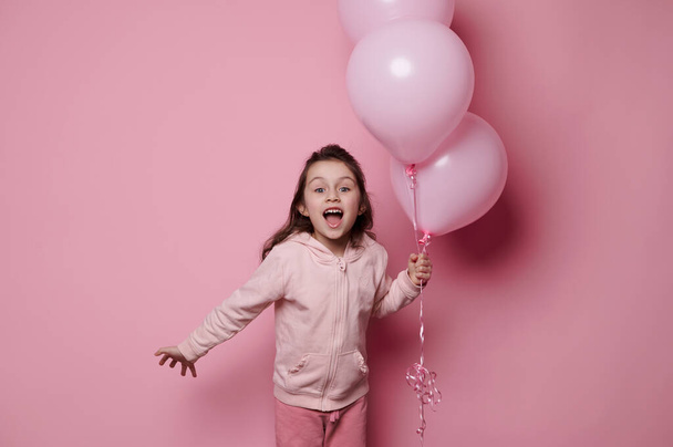 Isolated portrait on pink background of a happy amazed cheerful birthday girl in pink clothing, posing with a bunch of pink pastel helium balloons, expressing surprise and amazement, looking at camera - Photo, image