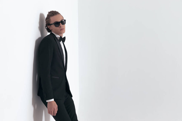 side view of blonde young groom with long hair wearing sunglasses and black tuxedo laying on a grey wall in studio - Foto, Imagem