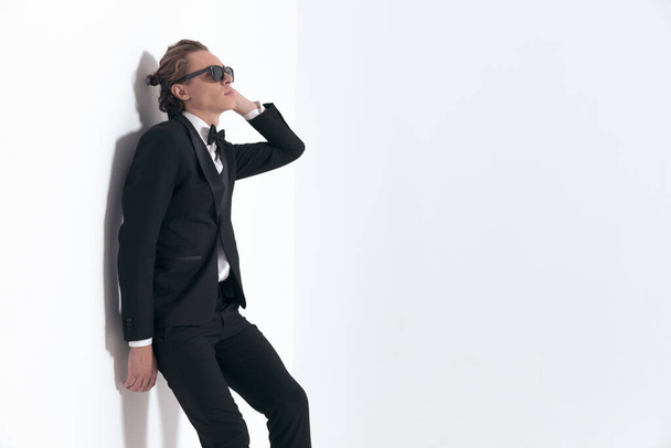 side view of attractive young man in tuxedo with sunglasses laying on a wall on grey background - Foto, Imagen