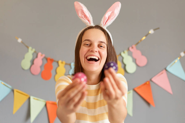 Portrait of laughing joyful woman wearing rabbit ears holding out two colorful Easter eggs, showing her hand painted eggs, being in good festive mood isolated on gray decorated background - Fotó, kép